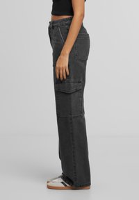 Pantalons cargo en denim gris foncé, avec une coupe décontractée, des poches latérales et une taille mi-haute. Associés à des baskets beiges.