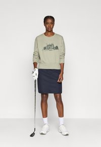Sweatshirt verde claro com um gráfico de castelo, acompanhado de uma saia azul marinha escura. Luva de golfe branca, sapatos e meias completam o visual.
