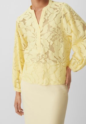 Blouse jaune à manches longues en dentelle avec motifs floraux, col en V, portée avec une jupe jaune clair, main posée sur la hanche.