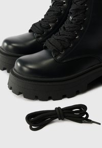 Czarne skórzane botki za kostkę z masywnymi gumowymi podeszwami, z teksturowanymi czarnymi sznurówkami i metalowymi oczkami. Zawiera dodatkową parę czarnych sznurówek.