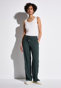 Grüne, strukturierte weit geschnittene Hosen mit einem Kordelzugbund, kombiniert mit einem weißen Tanktop und weißen Sneakers, vor einem schlichten Hintergrund.
