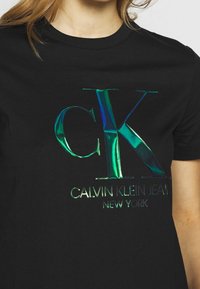 Černé bavlněné tričko s výrazným duhovým logem "CK" a textem "CALVIN KLEIN JEANS NEW YORK" pod ním v metalickém tisku.