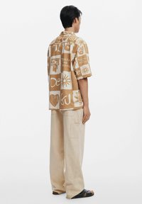 Kortærmet, beige skjorte med forskellige trykte designs i hvid, der har geometriske og abstrakte mønstre. Passer godt til lyse bukser.