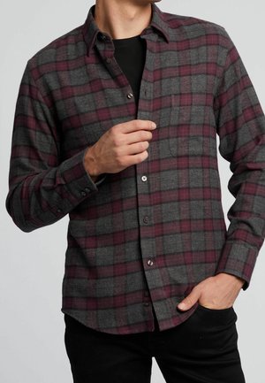 Homme portant une chemise à carreaux grise et bordeaux boutonnée sur un t-shirt noir, une main boutonnant la chemise et l'autre dans la poche.