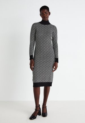 Abito in maglia con un motivo geometrico in bianco e nero, colletto alto, maniche lunghe e bordi a costine, abbinato a scarpe nere con tacco e cinta.