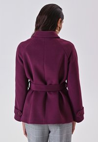 Cappotto viola con cintura e finitura testurizzata, dotato di colletto classico e polsini con bottone, indossato sopra pantaloni a scacchi. Vista posteriore mostrata.