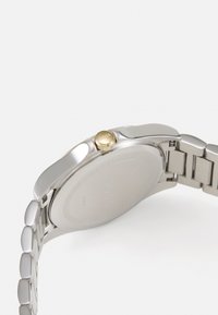 Montre en acier inoxydable argenté avec une finition polie, accent en or sur la couronne et un bracelet métallique épuré. Dos de boîtier rond visible.