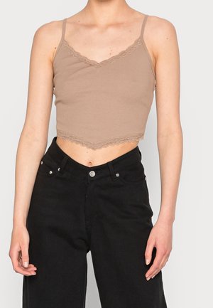 Femme portant un crop top beige avec bordure en dentelle et fines bretelles, et un pantalon noir taille haute, debout bras détendus.