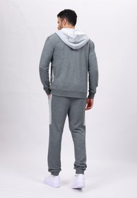 Grå treningssett som består av en hettegenser og matchende joggebukser, med lysere grå detaljer på sidene, og hvite sko.