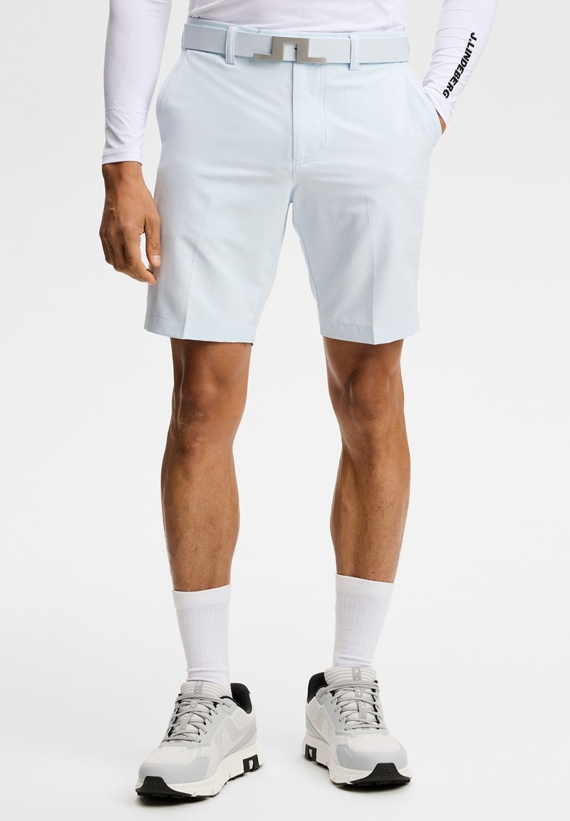 Man wearing light blue shorts, white belt, white long-sleeve top, white crew socks, and white athletic shoes standing with hands relaxed.