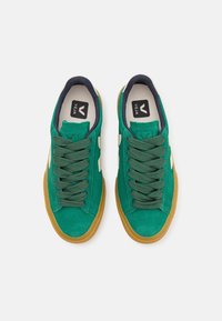 Gröna mockaskor med en gummisulor, utrustade med matchande skosnören och en vit logodetalj på sidan. Marinblå accenter runt öppningen.