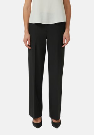 Kobieta ubrana w czarne spodnie z wysokim stanem, połączone z czarnymi szpilkami z zaostrzonymi noskami oraz białą, bezrękawnikową bluzką.