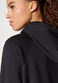 Μαύρη κουκούλα φούτερ με λεία υφή, διαθέτει μια διακριτική λογότυπο στην πλάτη, σχεδιασμένη με στενή εφαρμογή και αδιάκοπες λεπτομέρειες.