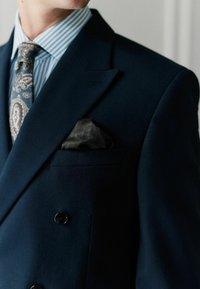 Tumes topeltpandidega ülikonnajakk, millel on teravad kraed; siidist tasku ruut, millel on mustriga tekstuur; sinise ja valge triibuline särk, paisley muster kaelarihm.