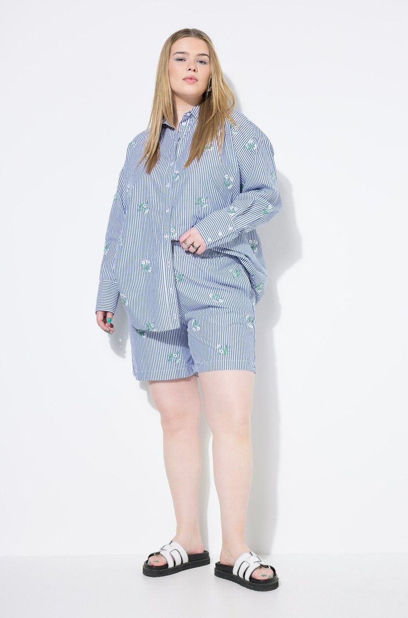 Conjunto de camiseta y pantalones cortos a rayas azules y blancas con bordados florales. La camiseta presenta mangas largas y un corte holgado. Sandalias deslizantes blancas.