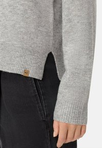 Pull en tricot rayé gris avec une fente sur le côté, présentant une finition texturée et une petite étiquette marron au bas. Jean en denim noir partiellement visible.