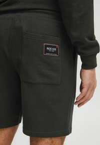 Mørkegrønne shorts lavet af bomuld, med en baglomme med et sort og rødt mærke. Inkluderer ribbede kanter og en afslappet pasform.