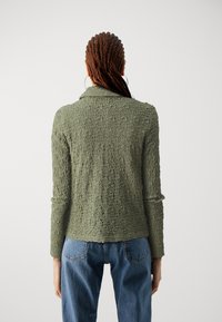 Suéter texturizado verde oliva con cuello y mangas largas, combinado con jeans de tiro alto azules, visto desde la espalda.