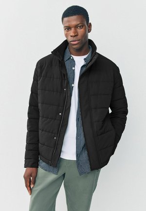 Chaqueta acolchada negra con un alto cuello, botones de presión plateados y una textura suave. Llevada sobre una camisa de cuadros azul y una camiseta blanca.