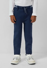Kind in marineblauwe broek met trekkoord en witte sneakers, staand tegen een effen lichtgrijze achtergrond.