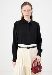 Chemise noire à manches longues, associée à une jupe crème plissée et à une fine ceinture noire. Tenant un petit sac à main rouge.
