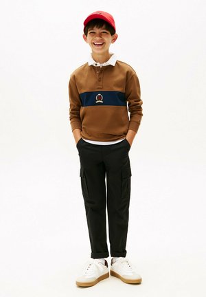 Polo à manches longues marron avec une rayure marine et un emblème, associé à un pantalon cargo noir et des baskets blanches avec des semelles beiges. Casquette rouge sur la tête.