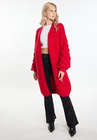Červený pletený kardigan s texturovanými širokými rukávy a žebrovaným designem. Nositý přes bílý crop top a spárovaný s černými rozšířenými kalhotami a špičatými botami.