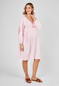 Rochie de maternitate roz cu mâneci lungi tip balon, decolteu în V, accente florale brodate, talie ajustată și fustă fluidă. Sandalele plate maro completează look-ul.