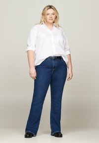 Modelka plus-size v bielej košeli s gombíkmi, modrých denimových zvonových džínsoch a čiernych topánkach. Košeľa má vyhrnuté rukávy, džínsy majú klasický strih.