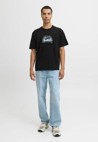 Czarna bawełniana koszulka z nadrukiem na piersi, w połączeniu z jasnoniebieskimi dżinsami. Model nosi beżowe i szare trampki.