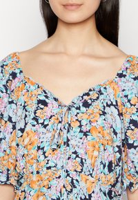 Blusa floral com mangas curtas em balão, apresentando um fundo azul-marinho, flores laranja e turquesa, e um detalhe de laço na gola.