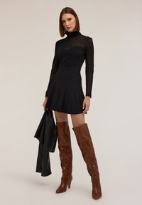 Černé plisované mini šaty s dlouhými krajkovými rukávy a stojáčkovým límcem, doplněné hnědými koženými vysokými botami a černou koženou bundou.
