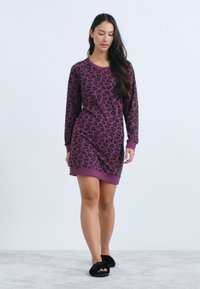 Vijolična obleka iz puloverja s črnimi leopardjimi vzorci, dolgimi rokavi in rebrastim robom. Model nosi črne poletne natikače na svetlem ozadju.