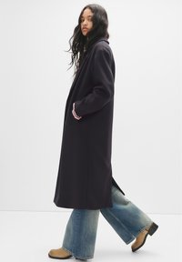 Donna con un lungo cappotto scuro, jeans ampi blu sbiaditi e stivali color crema che cammina di lato su uno sfondo bianco uniforme.