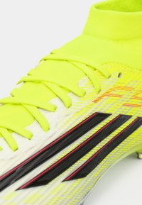 Λαμπερό κίτρινο ποδοσφαιρικό παπούτσι με υφή στο επάνω μέρος, μαύρες και κόκκινες διακοσμητικές ρίγες και σύστημα δέσης για σφιχτή εφαρμογή. Μεσαίου ύψους γιακά για υποστήριξη.