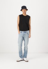Camiseta negra sin mangas, jeans azul claro y zapatillas blancas con acentos grises. Llevando un sombrero de cubo azul oscuro, de pie contra un fondo blanco.
