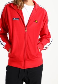 Rød lynlåstrøje i glat stof med hvide sidelister, en lille farverig logo og forlommer.