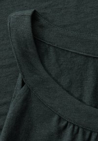 Tissu vert foncé au toucher doux ; présente un col rond et des fronces au niveau des coutures d'épaule, mettant en avant des éléments de design décontractés.