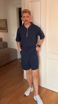 Jovem com cabelo loiro e bigode, vestindo uma camisa azul marinho com fecho, calções de igual cor e ténis brancos, a estar dentro de casa ao lado de uma porta branca.