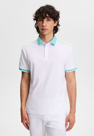 Ung mand med mørkt krøllet hår iført en hvid poloshirt med turkis krave og ærmekanter, stående mod en ensfarvet baggrund.