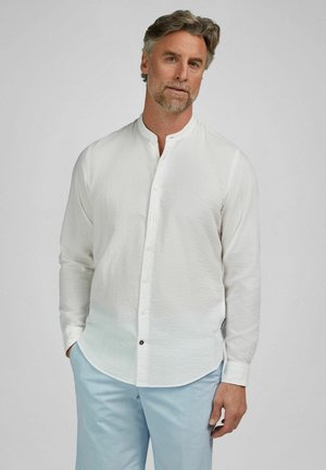 Man met grijs haar en baard, gekleed in een wit textuur overhemd met mandarijnkraag en lichtblauwe broek, staand tegen een effen grijze achtergrond.