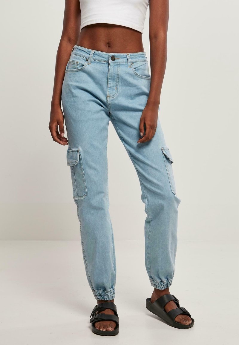 Femme portant un pantalon cargo en denim bleu clair avec des poignets élastiques, un débardeur court blanc et des sandales noires, debout devant un fond uni.