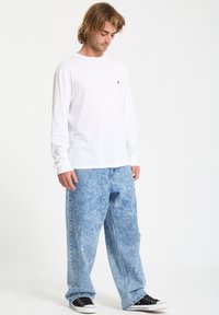 Weißes Langarm-T-Shirt mit kleinem schwarzem Logo, getragen mit hellblauen weiten Jeans mit einem strukturierten Finish und schwarzen Sneakers.