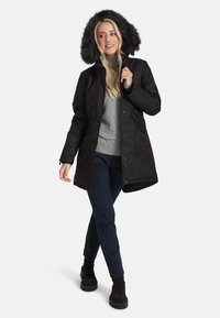 Czarna parka z kapturem wyłożonym sztucznym futrem, zamek błyskawiczny i zatrzaskowe zapięcie, średniej długości, zestawiona z szarym swetrem, granatowymi spodniami i czarnymi butami.