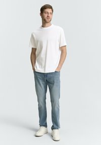 Biały bawełniany t-shirt z krótkim rękawem, w połączeniu z jasnoniebieskimi dżinsami i białymi trampkami. Model stoi z rękami w kieszeniach.
