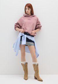 Felpa con cappuccio rosa, gonna di jeans con strisce blu legata in vita, stivali beige alla caviglia e calze bianche fin sopra il ginocchio. Look semplice e stratificato.