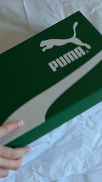 Caixa de sapatilhas verde da Puma com um logo branco e um acento prateado na tampa. Mão a segurar a caixa, colocada sobre papel branco amarrotado.