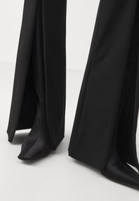 Zwarte broek met wijde pijpen en vooraan splitten die puntige zwarte laarzen onthullen, tegen een effen lichte achtergrond.