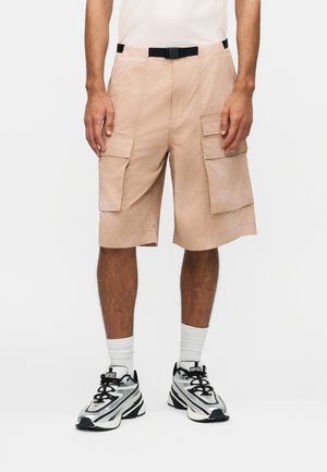 Άνδρας που φοράει μπεζ cargo σορτς με μεγάλες πλαϊνές τσέπες, μαύρη ζώνη, λευκές κάλτσες και ασπρόμαυρα αθλητικά παπούτσια, στέκεται μπροστά σε μονόχρωμο φόντο.