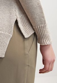 Beige gestrickter Pullover mit texturiertem, geripptem Saum und seitlichen Schlitzen, kombiniert mit glatten, hellbraunen Hosen, die eine lässige Passform zeigen.